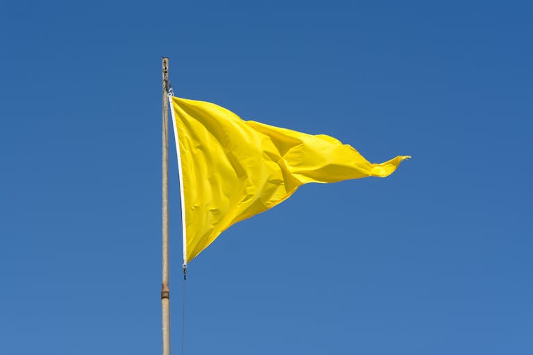 A vibrant yellow flag waving on a pole against a clear blue sky, conveying caution.