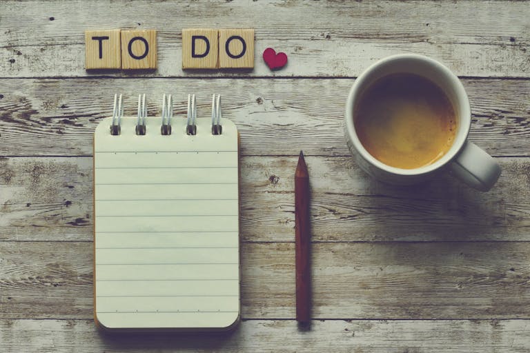 Flat lay of a notebook, pencil, coffee cup, and 'To Do' tiles on a rustic wooden table.
